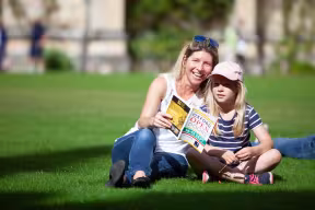 Woman and young girl sit on the grass reading Oxford Open Doors brochure