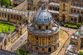 Aerial view of the Radcliffe Camera -a circular building topped with a dome- surrounded by historical university college buildings with enclosed grassy quads, and the University Church of St Mary the Virgin.