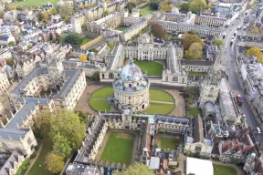 Aerial view of historical Oxford University buildings built in yellow stone, including All Souls College, the Bodleian Library and the Radcliffe Camera (a circular building with a dome). 