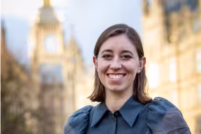 A young woman with shoulder length brown hair wearing a dark blue top. She stands outside with out of focus historical buildings in the background.
