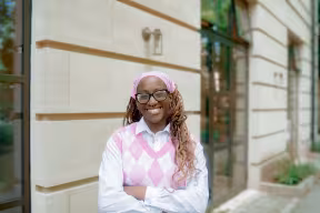 Seun Sowunmi wears a pink hairband, a white shirt, and a pink and white sleeveless jumper. She smiles with arms folded. She wears glasses, and her hair is in braids.