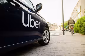 Side on view of a black car, with the brand name ‘Uber’ in white letters. A cyclist is visible ahead, in the background.