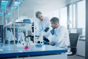 A man and woman wearing lab coats sit at a bench in a laboratory. In front of them, there is a microscope and a range of chemistry apparatus. 