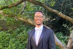 Jonathan Brooks wears academic dress, and a pair of glasses. He is smiling, standing in a garden, with a background of trees