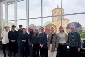 Students from the three winning projects stand together with Professor Irene Tracey in the Maths Institute. The Radcliffe Observatory can be seen in the background, through high windows.