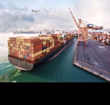 Aerial view of a large container ship docked at a port with cranes loading cargo, while an airplane flies overhead above the harbour.