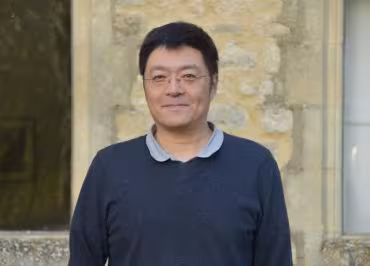 Outdoor portrait of a person wearing a dark sweater over a collared shirt, standing in front of a stone building wall in natural light.