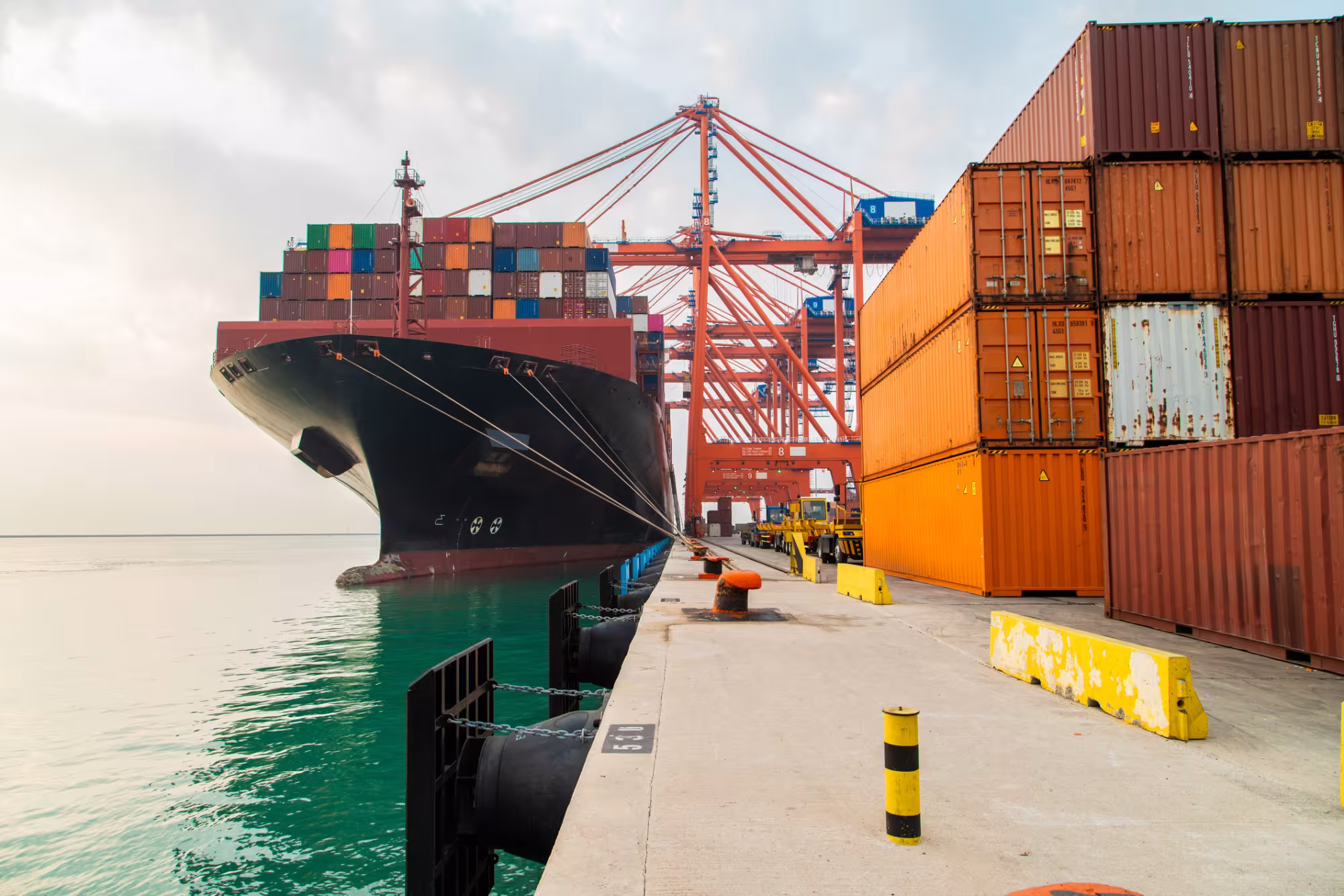 A cargo container ship moored at a harbour. Containers are being loaded or unloaded with cranes. 