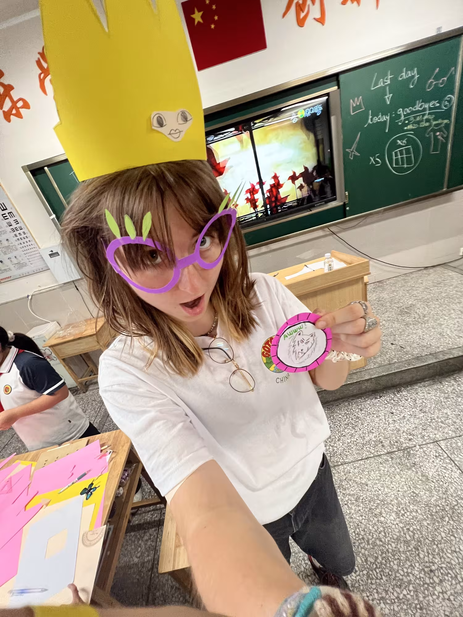 A student takes a selfie in a classroom, wearing oversized handmade purple glasses and a tall yellow paper hat. They hold up a colourful handmade badge featuring a drawing of a wolf. Craft materials, a chalkboard, a screen and a Chinese flag are visible in the background.
