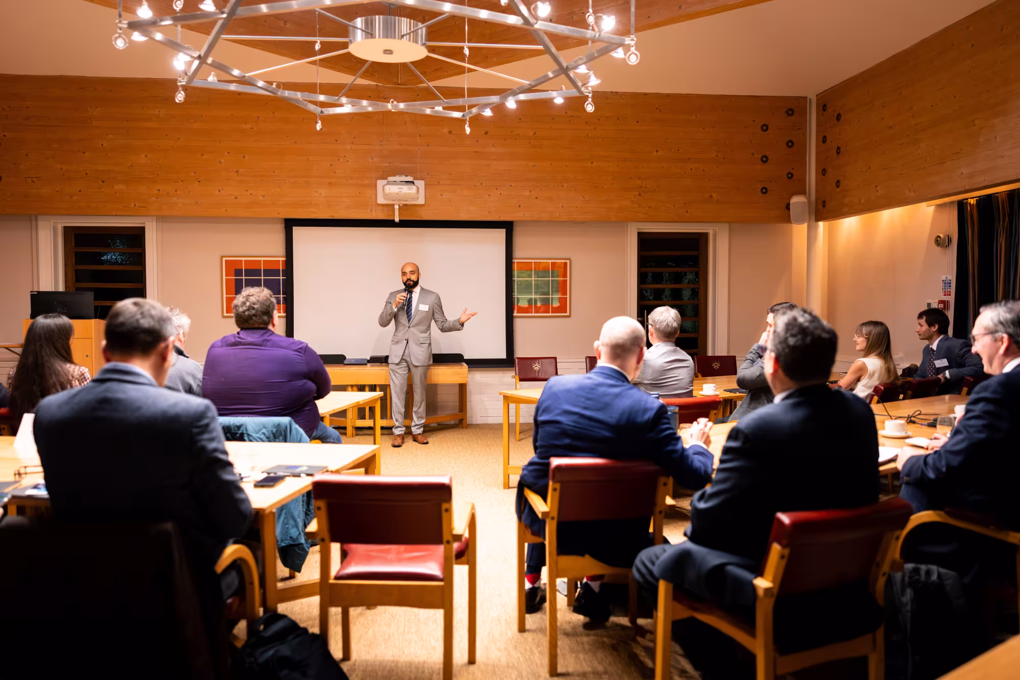 A speaker in a grey suit addresses an audience seated around tables in a conference room, holding a microphone and gesturing towards participants.